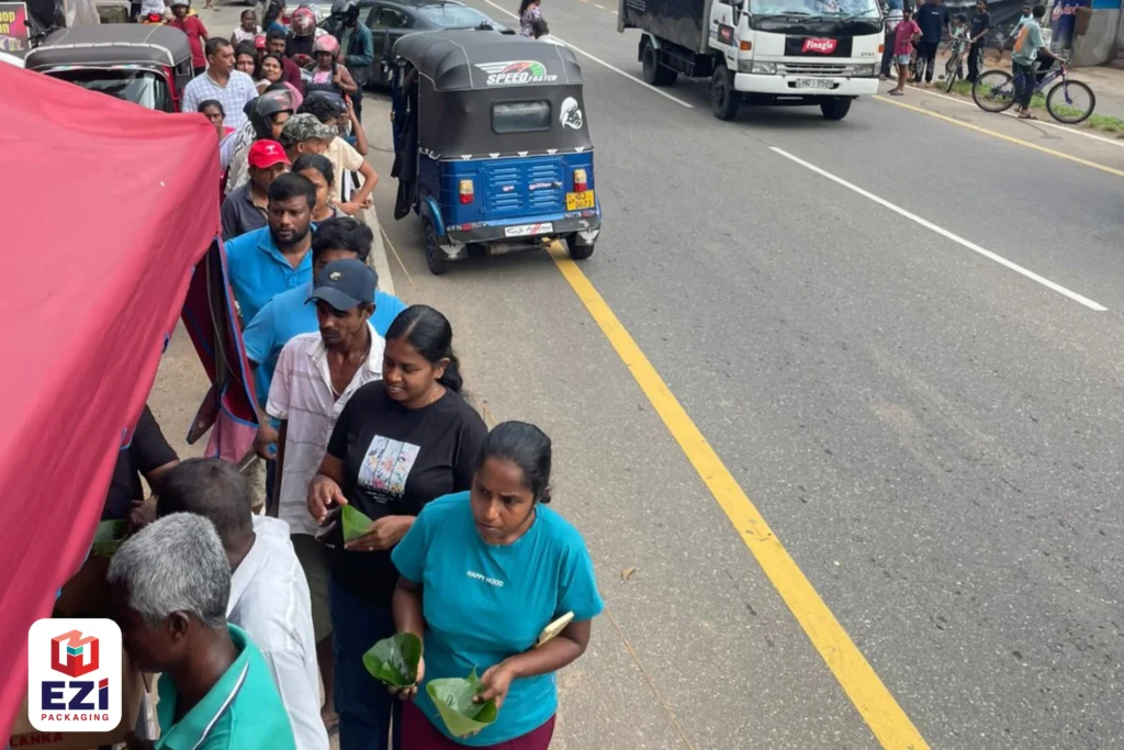 A long queue of local residents holding lotus leaves waiting alongside a road for the EZI Packaging dansala