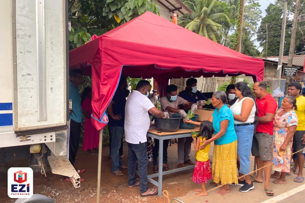 EZI Packaging team members serving food to community members under a red tent during a Vesak Poya Dansala event